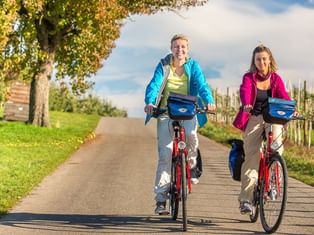 Two women cycling side by side on a paved path through autumn countryside with trees and vineyards under a blue sky.