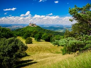 Burg Hohenzollern thront auf einem bewaldeten Hügel in Süddeutschland, umgeben von grünen Wiesen und Bäumen unter blauem Himmel mit weißen Wolken.