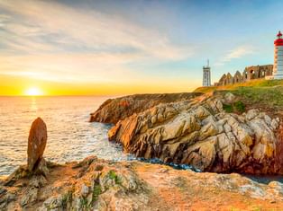 Rot-weiß gestreifter Leuchtturm in Plougonvelin auf Felsenklippen bei Sonnenuntergang, mit Ruinen und ruhigem Meer unter goldenem Himmel.