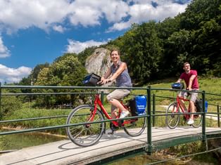 Zwei Radfahrer mit Packtaschen überqueren eine Holzbrücke mit grünem Geländer. Dichter Wald und Felsklippe im Hintergrund unter blauem Himmel.