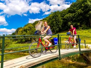 Zwei Radfahrer auf einer Metallbrücke über einen Bach bei Schelklingen an der Donau. Grüne Hügel und Felsen unter blauem Himmel.