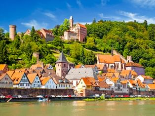 Historische Stadt Hirschhorn am Neckar mit Fachwerkhäusern, orangen Dächern und einer Burg auf dem Hügel unter blauem Himmel.