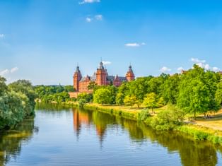 Schloss Johannisburg mit roten Sandsteintürmen spiegelt sich im Main, umgeben von grünen Bäumen unter blauem Himmel in Aschaffenburg.