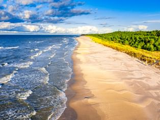 Luftaufnahme vom Strand Osieki mit sandigem Ufer, blauen Ostseewellen und dichtem grünem Wald unter teilweise bewölktem Himmel.