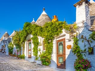 Reihe weißer Trulli-Häuser mit konischen Steindächern in Alberobello, geschmückt mit grünen Ranken und bunten Blumen unter blauem Himmel.