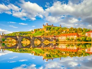 Festung Marienberg auf grünem Hügel über Würzburg mit Steinbrücke und bunten Häusern, die sich im Main unter blauem Himmel spiegeln.