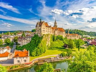 Hohenzollernschloss in Sigmaringen auf einem grünen Hügel über der Donau, umgeben von Bäumen und historischen Gebäuden unter blauem Himmel.