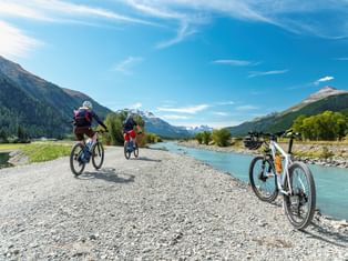 Zwei Radfahrer auf einem Schotterweg entlang des türkisfarbenen Inn-Flusses, mit einem geparkten Fahrrad im Vordergrund und Alpenbergen unter blauem Himmel.