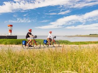 Zwei Radfahrer mit roten Helmen und blauen Taschen fahren entlang eines Weges am Wasser in Brunsbüttel. Ein rot-weißer Leuchtturm steht am Wasser.