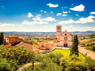 Basilika des Heiligen Franziskus in Assisi mit Glockenturm und grüner Rasenfläche, umgeben von Zypressen. Hügelige umbrische Landschaft.