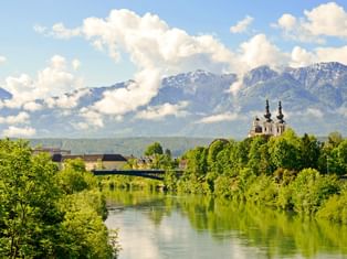 Blick auf Villach entlang der Drau mit grüner Vegetation, einer Kirche mit zwei Türmen und schneebedeckten Bergen unter bewölktem Himmel.