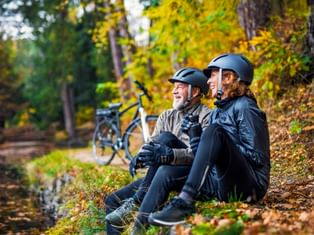 Zwei Radfahrer mit Helmen sitzen an einem Waldbach im Herbst, E-Bikes hinter ihnen geparkt. Buntes Herbstlaub umgibt die Szene.