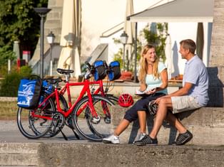 Zwei Radfahrer mit roten Tourenrädern rasten auf Steinstufen am Konstanzer Hafen. Historisches weißes Gebäude im Hintergrund, Räder mit Taschen.