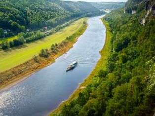 Luftaufnahme der Elbe bei Rathen, die sich durch grünes Tal mit bewaldeten Klippen schlängelt. Ein Raddampfer fährt auf dem ruhigen Wasser.