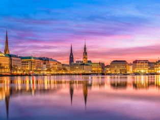 Hamburgs Binnenalster bei Sonnenuntergang mit beleuchteten historischen Gebäuden und Kirchtürmen, die sich im ruhigen Wasser unter rosa-blauem Himmel spiegeln.