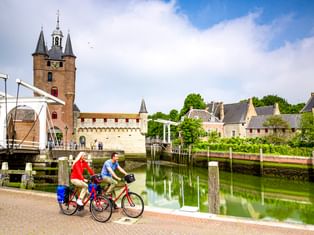 Two cyclists riding along a canal in Zierikzee with a white drawbridge, medieval tower, and historic buildings reflected in the water.