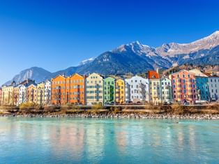 Reihe bunter historischer Gebäude entlang des türkisfarbenen Inn in Innsbruck mit schneebedeckten Berggipfeln im Hintergrund unter blauem Himmel.