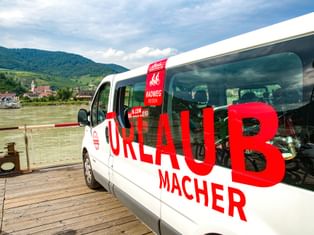 Weißer Radweg-Reisen Urlaubmacher Bus mit roter Beschriftung und Fahrrädern im Inneren, auf Holzsteg am Fluss mit Hügeln und Dorf im Hintergrund.