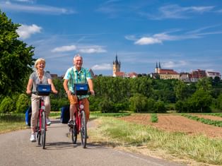 Zwei Radfahrer auf einem asphaltierten Weg durch Felder mit Bad Wimpfens historischer Skyline und Kirchtürmen im Hintergrund.