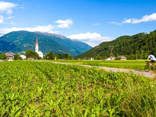 Radfahrer auf Weg durch grüne Felder bei Lienz in Osttirol, mit Kirchturm und Dolomiten im Hintergrund unter blauem Himmel.