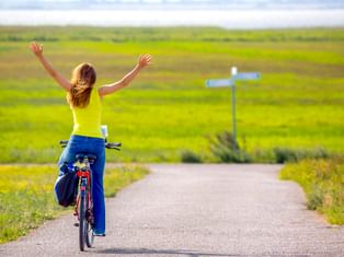 Frau auf Fahrrad mit freudig erhobenen Armen auf Weg durch grüne Wiesen in Ahrenshoop. Ein weißer Wegweiser steht neben dem Weg.