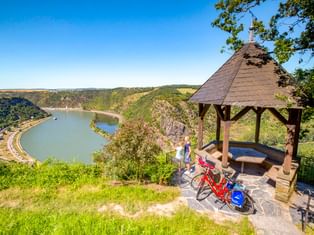 Holzpavillon am Aussichtspunkt Maria Ruh mit rotem Fahrrad, Blick auf die Rheinschleife bei Loreley mit grünen Hügeln und blauem Himmel.