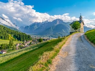 Kleine Kapelle mit Glockenturm auf grünem Hügel bei Toblach, Südtirol. Ein Schotterweg führt zur Kapelle mit Berggipfeln im Hintergrund.
