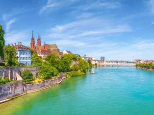 Türkisfarbener Rhein fließt durch Basel mit historischen Gebäuden, roten Kirchtürmen und Steinmauer unter blauem Himmel.