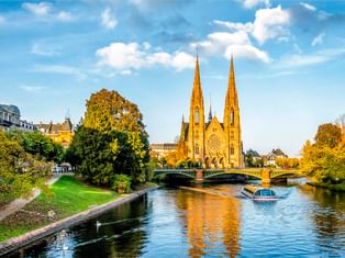 Gotische Kirche mit zwei Türmen in Straßburg neben einem Kanal mit Boot. Grüner Park und Brücke im Vordergrund unter blauem Himmel mit Wolken.