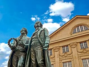 Bronzenes Goethe-Schiller-Denkmal in Weimar mit zwei stehenden Figuren vor blauem Himmel und klassizistischem Theatergebäude mit Rundbogenfenster.