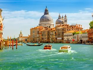 Boote auf dem Canale Grande in Venedig mit historischen Gebäuden und der Kuppelbasilika Santa Maria della Salute unter blauem Himmel.