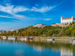 Bratislavaer Burg auf einem grünen Hügel mit Blick auf die Donau mit Kreuzfahrtschiffen unter blauem Himmel mit weißen Wolken.
