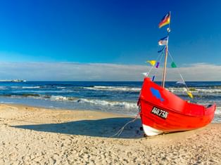 Rotes Fischerboot mit bunten Flaggen am Sandstrand in Ahlbeck, Usedom. Blaue Ostsee mit sanften Wellen unter klarem Himmel.