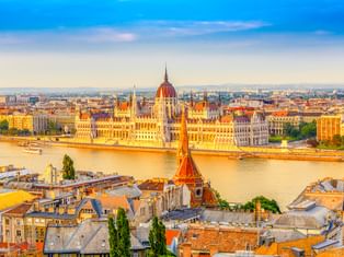 Luftaufnahme des ungarischen Parlamentsgebäudes an der Donau in Budapest, mit historischen Gebäuden und Dächern im Vordergrund.