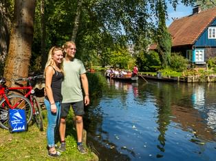 Paar mit Fahrrädern am Kanal im Spreewald. Traditionelles blaues Haus mit rotem Dach und Touristenboot im Hintergrund sichtbar.