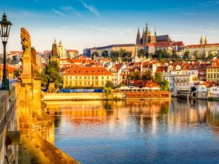Blick von der Karlsbrücke in Prag mit Statue und Laterne, Moldau, historischen Gebäuden mit roten Dächern und Prager Burg auf dem Hügel.