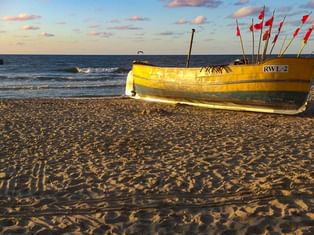 Gelbes Fischerboot mit Kennzeichen RWL-2 und roten Flaggen am Sandstrand bei Sonnenuntergang. Ostseewellen und blauer Himmel mit rosa Wolken im Hintergrund.