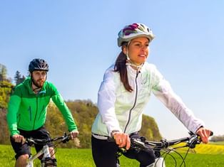 Zwei Radfahrer fahren durch grüne Landschaft am Bodensee. Frau in weißer Jacke führt, Mann in grüner Jacke folgt an sonnigem Tag.