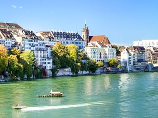 Rhein mit grünem Wasser fließt an Basels historischer Uferpromenade vorbei. Bunte Gebäude und ein Kirchturm säumen das Flussufer mit Bäumen und Booten.