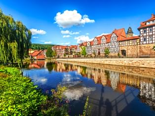 Historische Fachwerkhäuser am Ufer in Hann. Münden spiegeln sich im ruhigen Flusswasser, mit Steinmauer und Weide im Vordergrund.