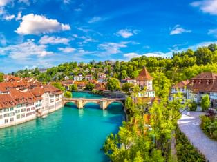 Nydeggbrücke über die türkisfarbene Aare in Bern mit historischen Gebäuden, roten Ziegeldächern, Kirchturm und grünem Hang unter blauem Himmel.