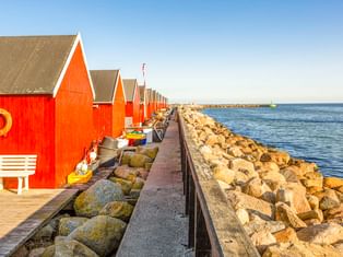 Reihe roter Holzbootshäuser mit weißer Verzierung entlang eines Steinpiers im Hafen von Rødvig. Kleine Boote neben den Gebäuden, ruhige blaue See.