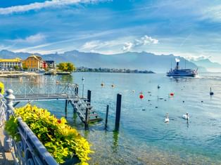 Seepromenade in Vevey mit Bänken, Blumen und Schwänen auf dem Genfer See. Berge und ein Raddampfer im Hintergrund sichtbar.