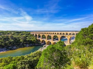 Pont du Gard, eine dreistöckige römische Aquäduktbrücke bei Remoulins, die einen Fluss überspannt, umgeben von üppiger grüner Vegetation unter blauem Himmel.