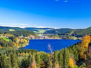 Blauer See umgeben von bewaldeten Hügeln im Schwarzwald. Herbstbäume mit gelbem und orangem Laub rahmen die Aussicht unter blauem Himmel.