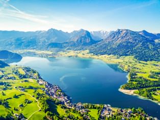 Luftaufnahme vom Wolfgangsee umgeben von grünen Wiesen und Bergen. Kleine Dörfer säumen das Ufer, mit Alpengipfeln im Hintergrund.