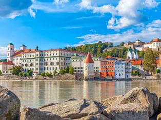 Bunte historische Gebäude der Passauer Altstadt an der Donau mit weißem Turm mit rotem Dach, vom felsigen Flussufer unter blauem Himmel.