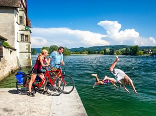 Zwei Radfahrer mit Rädern pausieren an einem See, während zwei Personen ins Wasser springen. Historisches Gebäude links, grüne Hügel im Hintergrund.