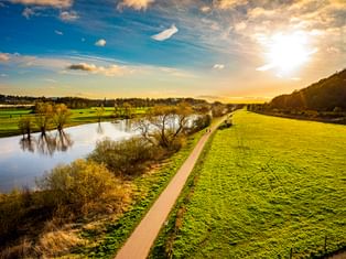 Asphaltierter Radweg entlang der Weser bei Porta Westfalica bei Sonnenuntergang. Grüne Wiesen säumen den Weg, Bäume stehen im Wasser, Hügel im Hintergrund.