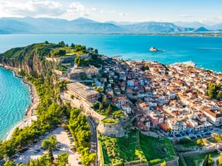 Luftaufnahme der Halbinsel Nafplio mit historischer Festung, rotgedeckten Gebäuden und türkisfarbener Bucht. Berge im Hintergrund unter blauem Himmel.